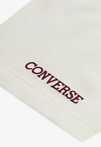 Beige bomullsstoff med synlig tekstur. "CONVERSE" trykt i burgunder nær kanten, som fremhever merkevaren.