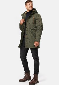 Olivgrüne Parka-Jacke mit Kapuze, ausgestattet mit Reißverschluss und Knopfverschluss, seitlichen Taschen und strukturiertem Stoff. Getragen über dunklen Jeans und braunen Stiefeln.
