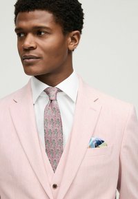Ljust rosa kostym med notch-lapel, vit skjorta och mönstrad rosa slips. Bröstficka med blå, grön och orange detalj. Texturerad tyg.