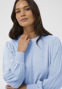 Hele sinine kootud kampsun soonikkoes, laienenud varrukatega. Ümar kaelus ja tekstuuriga kangas. Lähedalt vaadatuna naine, kes õrnalt enda kaela puudutab.