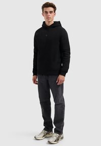 Jonge man die staat en een zwarte hoodie, donkere spijkerbroek en witte sportschoenen draagt tegen een effen lichte achtergrond.