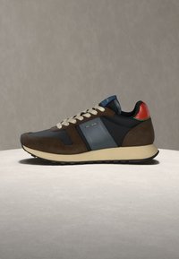Baskets marron, bleu marine et noires avec des lacets crème, une semelle beige et un détail rouge au talon sur une surface claire sur un fond neutre.