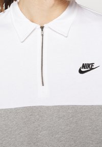 Nike pikétröja med en vit överdel och en grå underdel, kvartsglasöppning och svart broderad logotyp på bröstet.