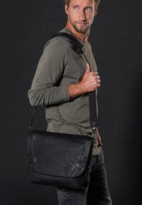 Zwarte leren boodschappentas met klepsluiting, verstelbare riem en geperst logo. Soepele textuur met minimale hardware-accenten.