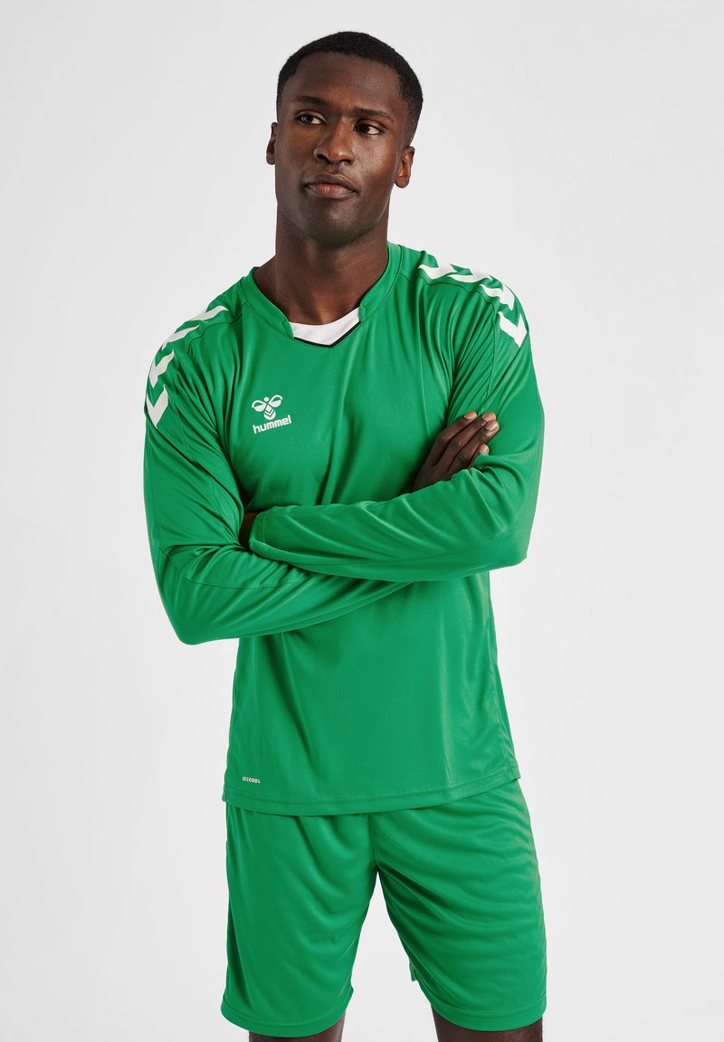 Man in groen voetbaltshirt met lange mouwen en korte broek met wit logo en chevronstrepen, staat met gekruiste armen en kijkt opzij.