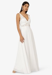 Weißes Kleid mit Spitzenverzierungen am Oberteil, einem tiefen V-Ausschnitt und einem fließenden Rock, kombiniert mit silbernen Ohrringen und Riemchensandalen.