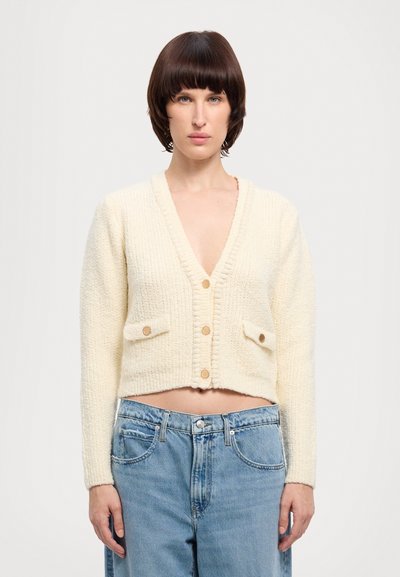 Femme aux cheveux bruns courts portant un cardigan crème boutonné avec poches avant et un jean taille haute bleu clair, sur un fond uni.