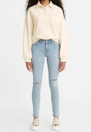 Kobieta w kremowej, długorękawowej koszulce polo, jasnoniebieskich, podartych obcisłych dżinsach oraz białych trampkach z panterkowymi detalami.