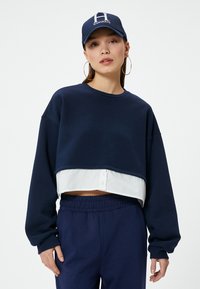 Sudadera navy recortada con una camisa blanca debajo, con mangas largas y cintura elástica, combinada con una gorra a juego.
