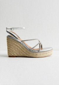 Silver wedge sandal with braided jute outsole, metallic straps, and ankle buckle. Smooth toe area and decorative accents along the straps.