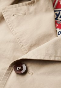 Beige jasstof met een soepele textuur; voorzien van een donkerbruine knop en zichtbare stiknaaddetails. Binnenvoering met een rood en blauw patroon.