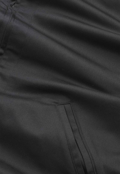 Primo piano di tessuto nero con cucitura visibile e dettaglio tasca a filetto, che mostra una texture liscia e pieghe sottili.