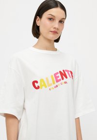 Valkoinen puuvillapaita rentoa istuvuutta, jossa on rohkeat punaiset, keltaiset ja vaaleanpunaiset graafiset tekstit, jotka sanovat "CALIENTE IT'S NOW COOL."