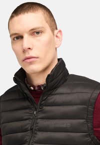 Vest puffer negru cu guler înalt, cusături orizontale și fermoar lateral. Prezintă un logo discret Timberland pe piept.