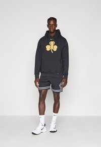 Felpa nera con un grande logo dorato a forma di trifoglio, abbinata a pantaloni corti in rete neri con strisce bianche e scarpe da ginnastica bianche.