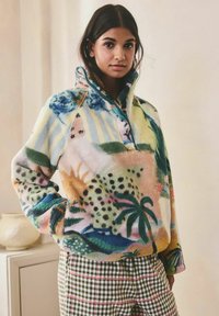 Femeie cu păr închis la culoare purtând un pulover fleece cu model colorat și pantaloni cu carouri verzi, stând în interior lângă un dulap deschis cu o vază.