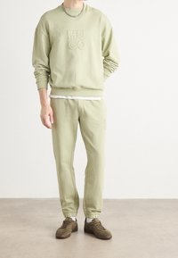 Persoon draagt een lichtgroene HUGO-sweatshirt en bijpassende joggingbroek met bruine sneakers, staand op een effen vloer tegen een witte muur.