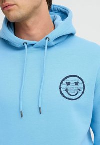 Felpa blu chiaro con cappuccio, caratterizzata da una grafica rotonda con testo e una faccina sorridente sul lato sinistro del petto, con cordini e terminali in metallo.