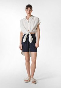 Vitrandig skjorta med knappar knuten i midjan, kombinerad med marinblå shorts och beige sandaler. Utseendet är avslappnat och ledigt.