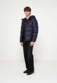 Marineblaue Steppjacke mit durchgehendem Reißverschluss, Kapuze und Logo auf der Brust; kombiniert mit schwarzer Hose und Schuhen, die eine lässige, figurbetonte Silhouette zeigen.