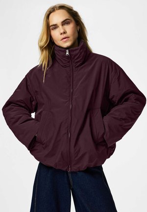 Personne aux longs cheveux blonds portant une doudoune violet foncé, les mains dans les poches, et un jean bleu foncé sur un fond uni.