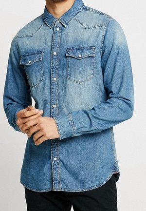 Mann trägt ein hellblaues Jeanshemd mit zwei Brusttaschen und schwarze Hose, Hände vor dem Körper verschränkt.