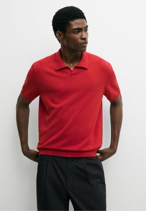 Homme portant un polo rouge à manches courtes et un pantalon noir, debout les mains sur les hanches, regardant sur le côté sur un fond uni.