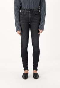 Fekete, skinny farmernadrág, magas derekú fazonnal, enyhén textúrált denim anyagból, párosítva egy bordázott sötétkéks pulóverrel és fekete lapos cipővel.