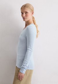 Lichtblauwe langetermijnsleeve shirt van zachte, gestructureerde stof met een ronde halslijn en subtiele branding op de borst. Gecombineerd met beige broek.