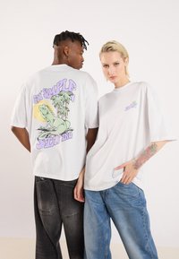 Άσπρες υπερμεγέθεις μπλούζες τύπου t-shirt; Η πλάτη έχει πολύχρωμο γραφικό με έναν σαύρα και φοίνικες. Το μπροστά μέρος έχει μικρό λογότυπο σε παστέλ χρώματα. Άνετη εφαρμογή.