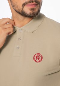 Beige polo tričko z texturované látky s třemi knoflíky, žebrovaným límečkem a červenou vyšitou emblémem na hrudi.