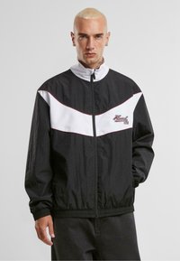 Veste coupe-vent noir et blanc avec un col montant, fermeture éclair, motif en chevrons et logo brodé en rouge. Tissu texturé.