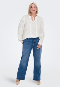 White long-sleeve blouse with decorative stitching and a tie neck, paired with high-waisted blue wide-leg jeans and beige heels.