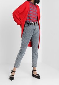 Röd ribbade kofta över en röd och blå rutig skjorta, i kombination med högmidjade ljusblå jeans och svarta klackskor.