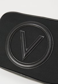 Μαύρη τσάντα με υφή, που φέρει αναγλυφό κυκλικό λογότυπο με στυλιζαρισμένο "V", κατασκευασμένη από υλικό που μοιάζει με δέρμα. Απλός, μοντέρνος σχεδιασμός.