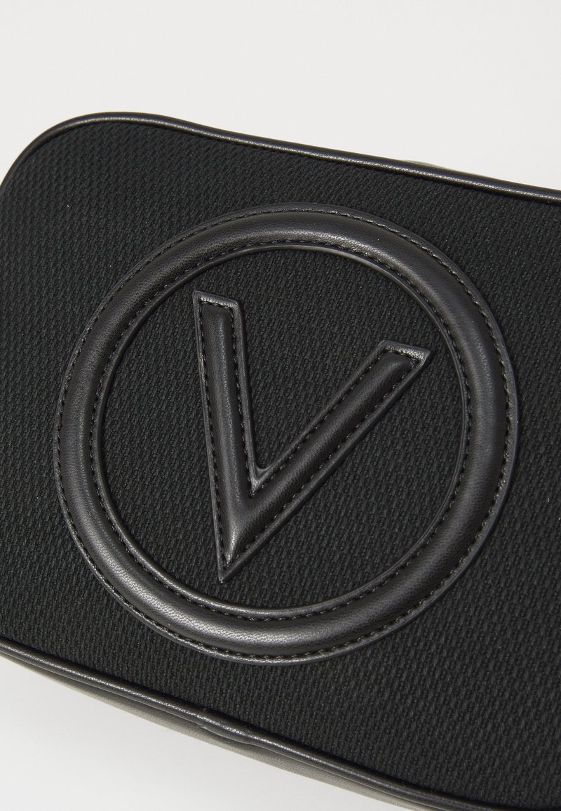 Sac noir texturé avec un logo circulaire en relief portant un « V » stylisé, fabriqué en matériau lisse semblable au cuir. Design simple et moderne.