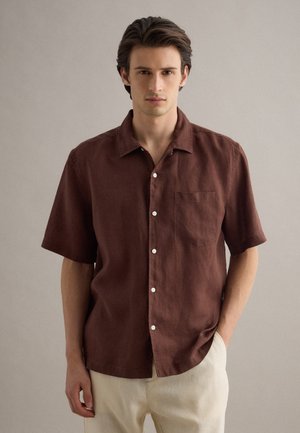 Giovane uomo che indossa una camicia marrone a maniche corte con taschino frontale e pantaloni beige chiaro su uno sfondo neutro e uniforme.