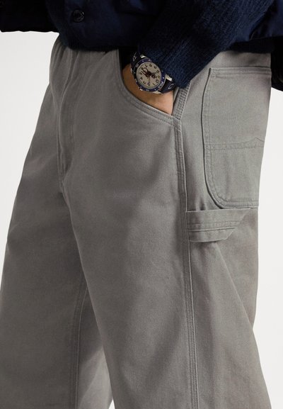 Harmaat puuvillahousut rentolla istuvuudella, joissa on sivutaskut ja näkyvät saumat. Ranteessa on sininen kello.