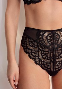 Melni mežģīņu augsta vidukļa biksītes ar sarežģītiem viļņotiem rakstiem, caurspīdīgiem paneļiem un gludu audumu, kontrastējošas faktūras vidukļa rajonā.