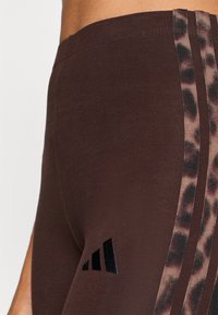 Καφέ αθλητικό κολάν με ελαστική μέση, με ρίγες λεοπάρ στο πλάι και μαύρες λογότυπες λεπτομέρειες στο κάτω μέρος του ποδιού.