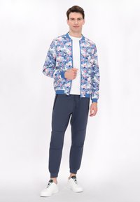 Giacca bomber floreale con sfondo blu, caratterizzata da motivi rosa e verdi. Abbinata a pantaloni jogger blu navy e sneakers bianche.