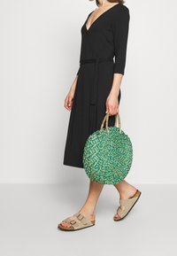 Svart omlottklänning med trekvartsärmar, tillsammans med en rund grön och naturlig stråväska och beiga sandaler med spännen.