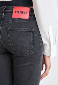 Primer plano de un bolsillo de vaqueros negros con una etiqueta roja de la marca HUGO, combinado con una camisa blanca de manga larga en una persona de pie.