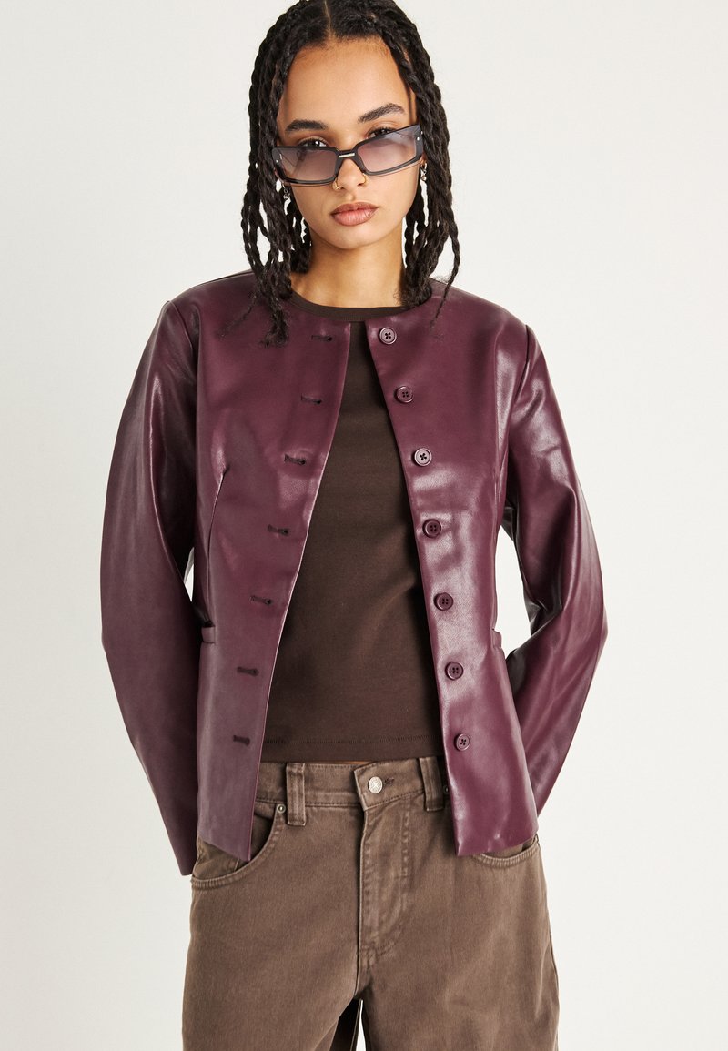 Fialová kožená bunda s okrúhlym výstrihom, predným zapínaním na gombíky a dlhými rukávmi. Skombinovaná s hnedým topom a tmavohnedými nohavicami.
