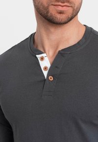 Lähikuva miehestä, joka käyttää tummanharmaata henley-paitaa, jossa on kolme ruskeaa nappia ja valkoista kangasta kauluksen sisäpuolella.