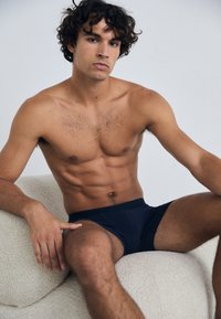 Marineblå truser på en skjorteløs mannemodell med krøllete hår, sittende mot en lys, teksturert bakgrunn. Definerte magemuskler og muskuløse armer synlige.