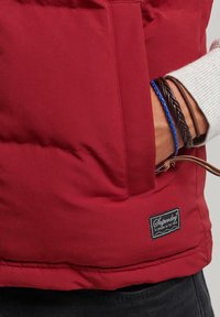 Doudoune rouge avec une texture lisse, comportant une poche à patte et une étiquette de marque. Accessoirisée avec des bracelets tressés de différentes couleurs.
