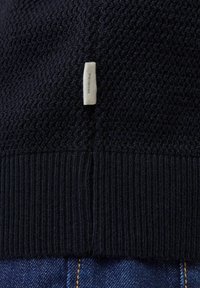Nahaufnahme eines dunklen Strickstoffs mit einem kleinen weißen rechteckigen Etikett in der Mitte über dem gerippten Saum, liegend auf blauem Denim mit sichtbaren Nähten.