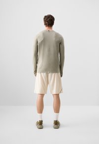 Langærmet olivengrøn skjorte, lysecreme shorts, atletiske sko, synlige stikninger og tekstureret stof. Bagvinkel.