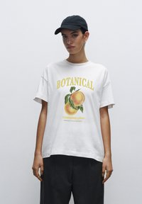 Bílý bavlněný tričko s barevným potiskem švestky a žlutým textem "BOTANICAL Champion plum" v uvolněném střihu. Nosená černá kšiltovka.
